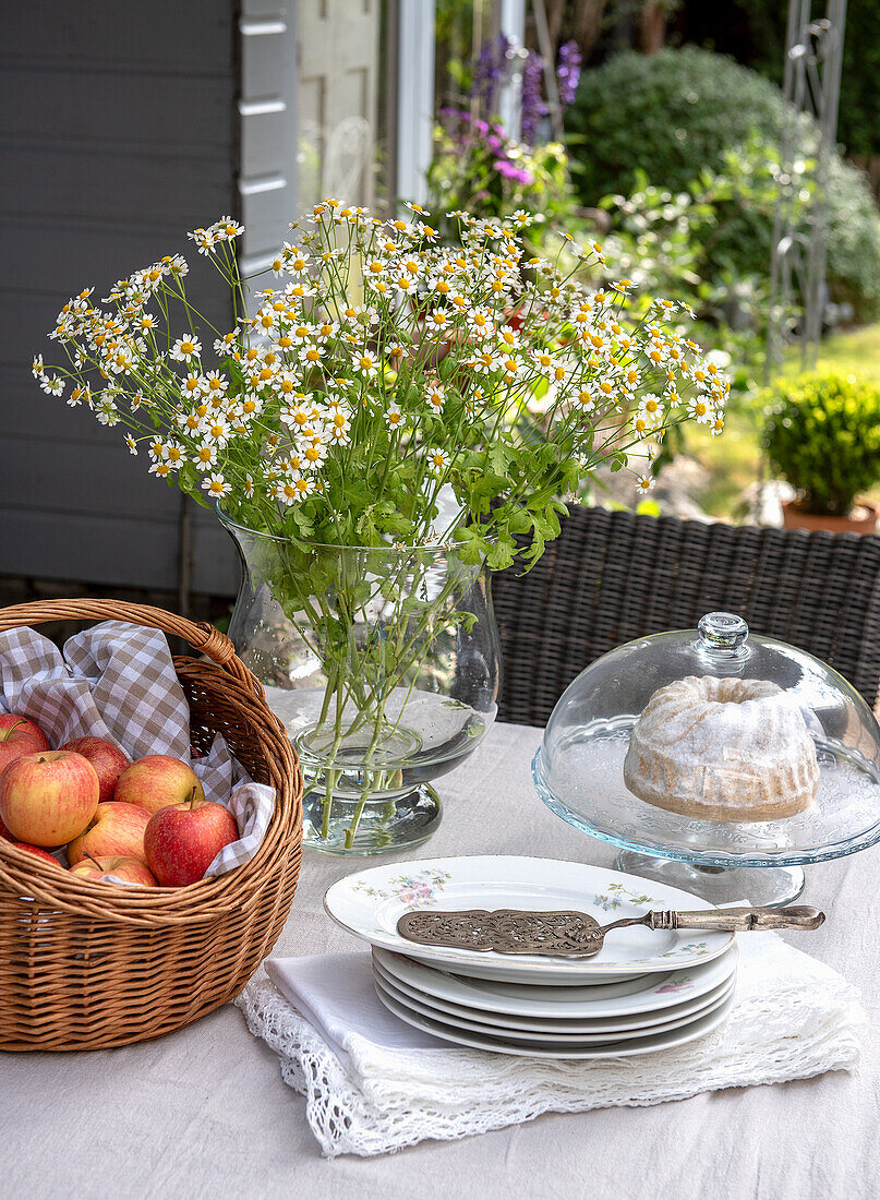 Gedeckter Gartentisch mit Apfelkorb, Gugelhupf und Gänseblümchenstrauß