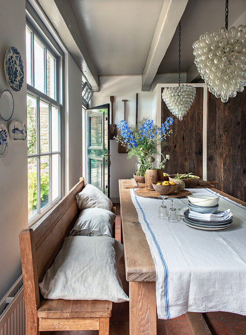 Rustic dining area with wooden table and bouquet of flowers, bench with linen cushions, glass bead chandelier