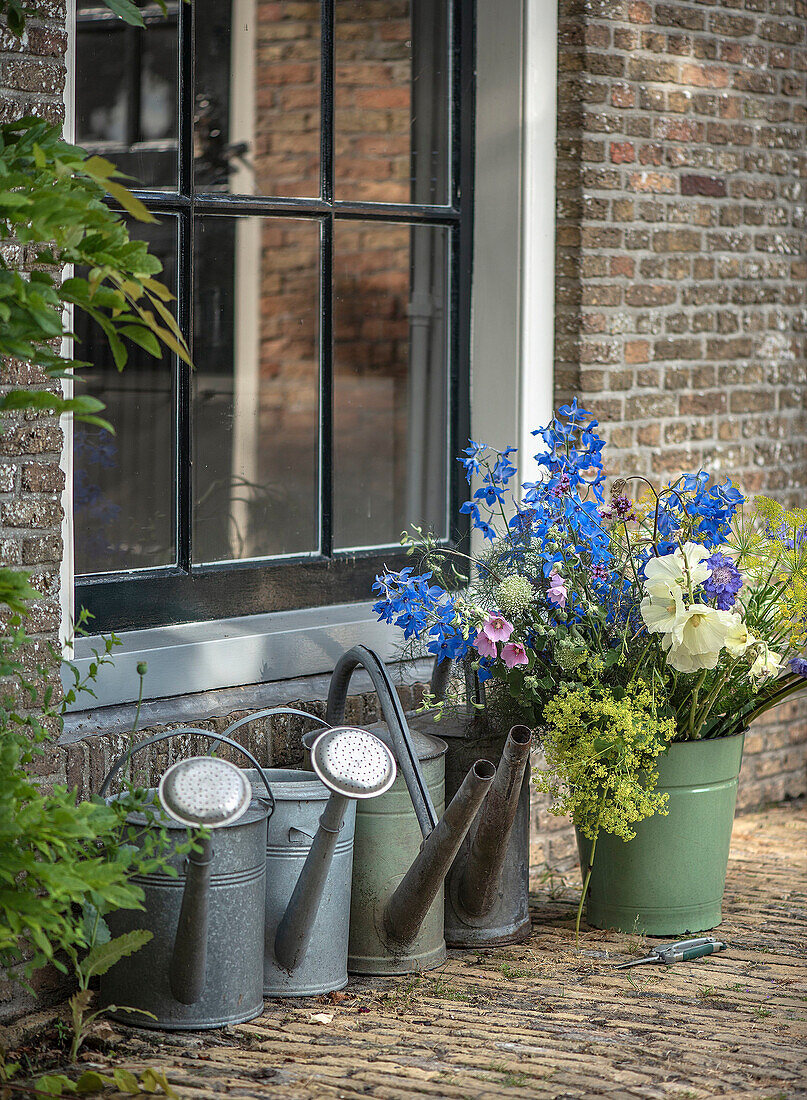 Several watering cans and colorful bouquet of flowers in front of brick house