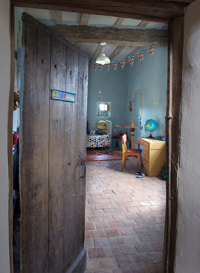 Blick durch alte Holztür in Kinderzimmer mit Terrakottafliesenboden und rustikalem Holzbalkendecke