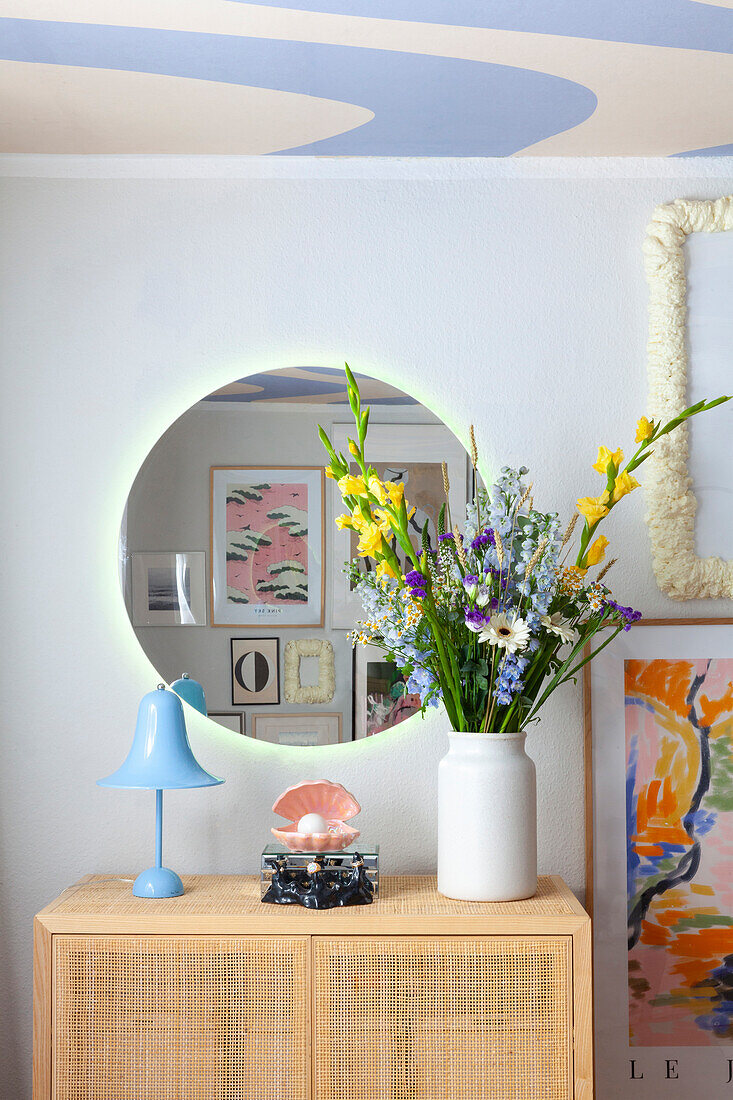 Bouquet of flowers in white vase on rattan dresser, blue table lamp and round mirror on the wall