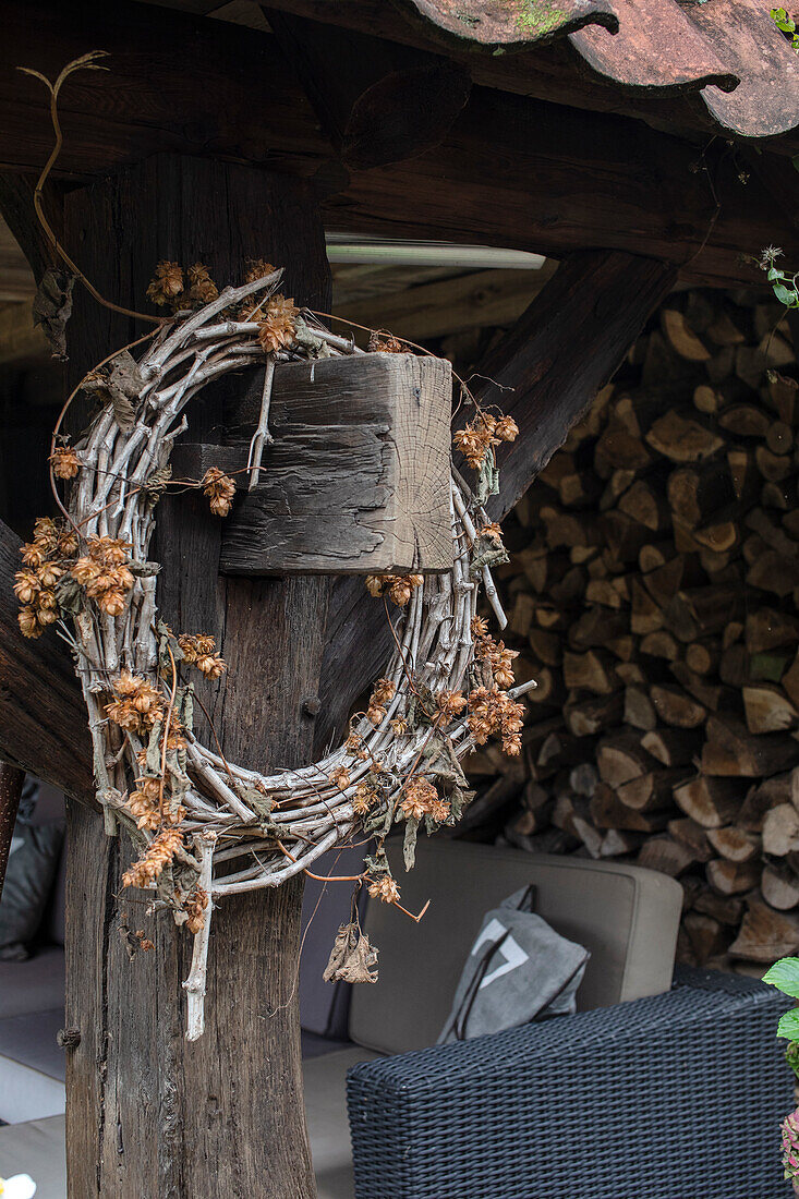 Rustic wreath on wooden pillar next to stacked firewood and rattan sofa