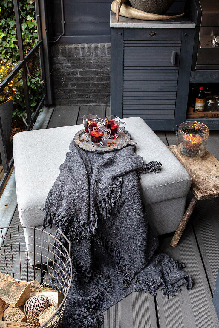 Grey-covered pouf with woollen plaid, tray with mulled wine and basket with firewood on the patio