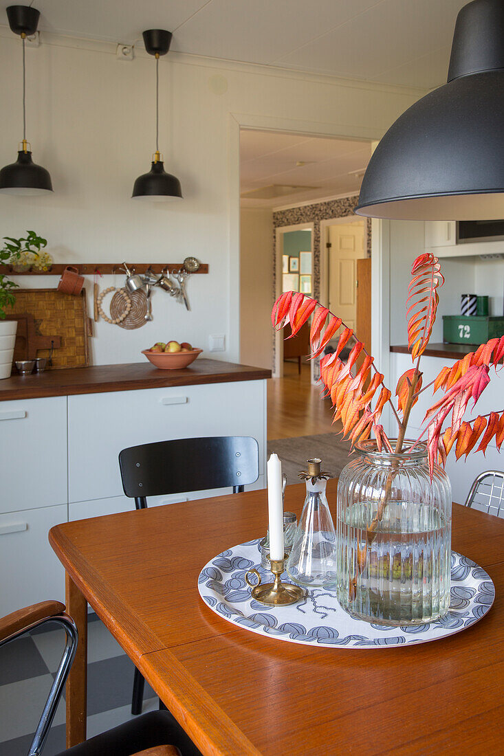 Glass vase with red-orange colored leaves on wooden table in kitchen