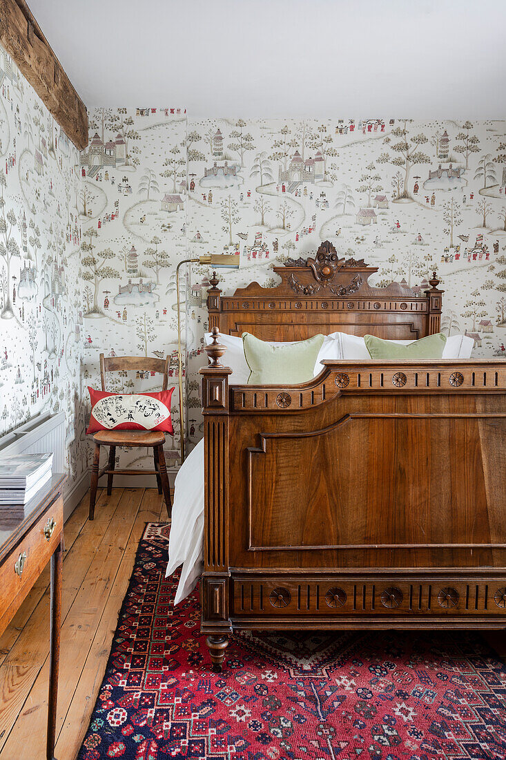 Holzbett mit Schnitzereien und gemusterte Tapete im Schlafzimmer mit Teppich und Holzdielenboden