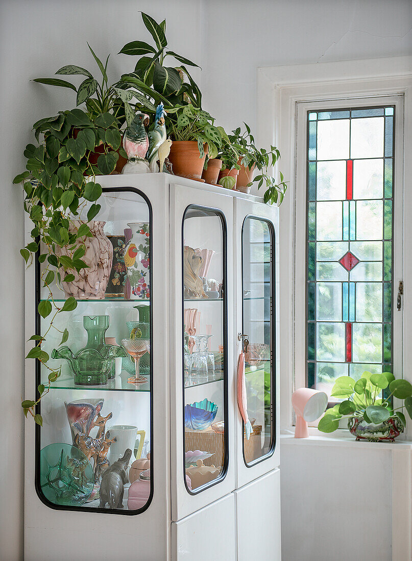Vitrinenschrank mit Pflanzen, Vintage-Geschirr und Vasen neben Buntglasfenster