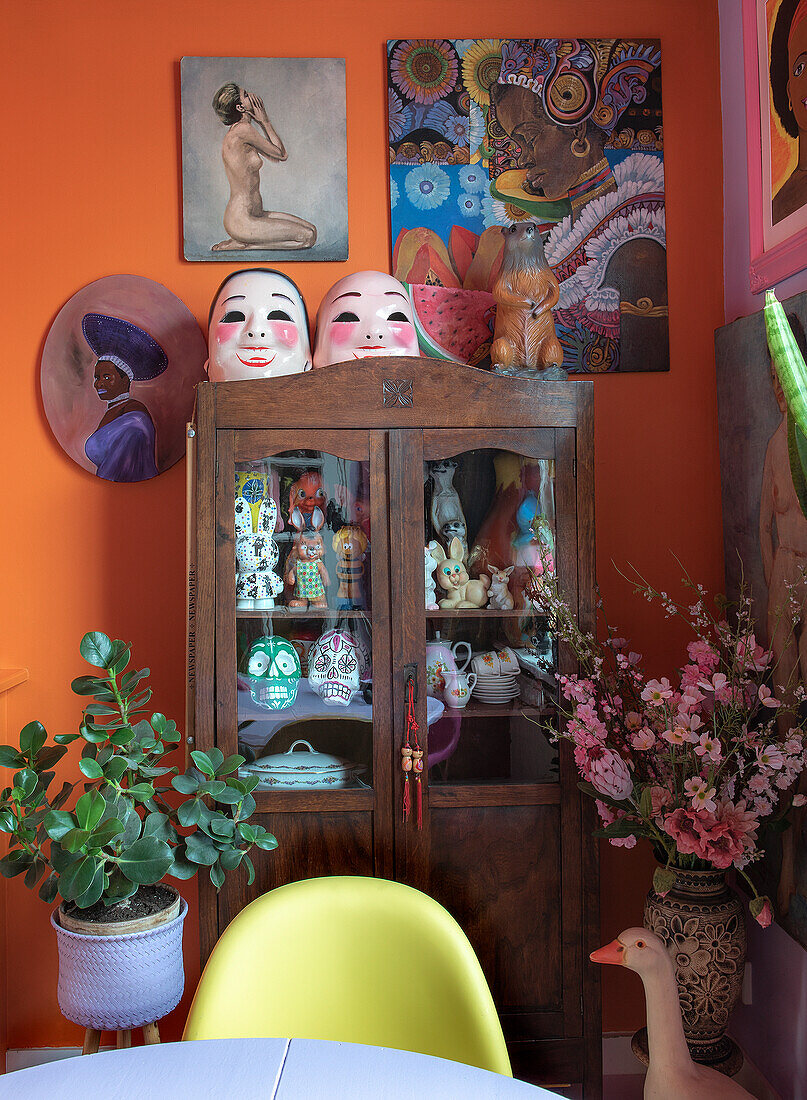 Display cabinet with figurines and masks, surrounded by colorful artwork and plants