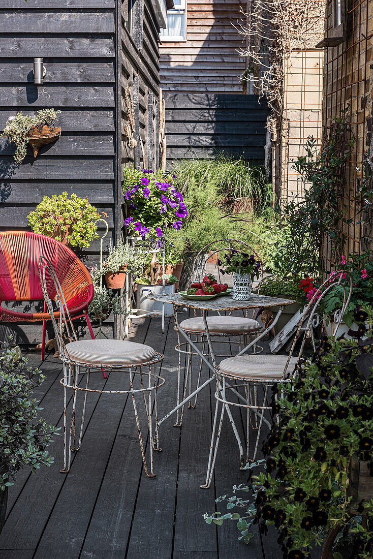 Small seating area with metal chairs and table on a wooden terrace, surrounded by flowering plants