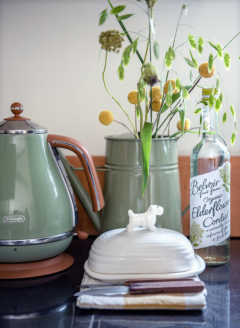 Green teapot, butter dish and elderflower syrup next to flower arrangement of yellow ball blossoms