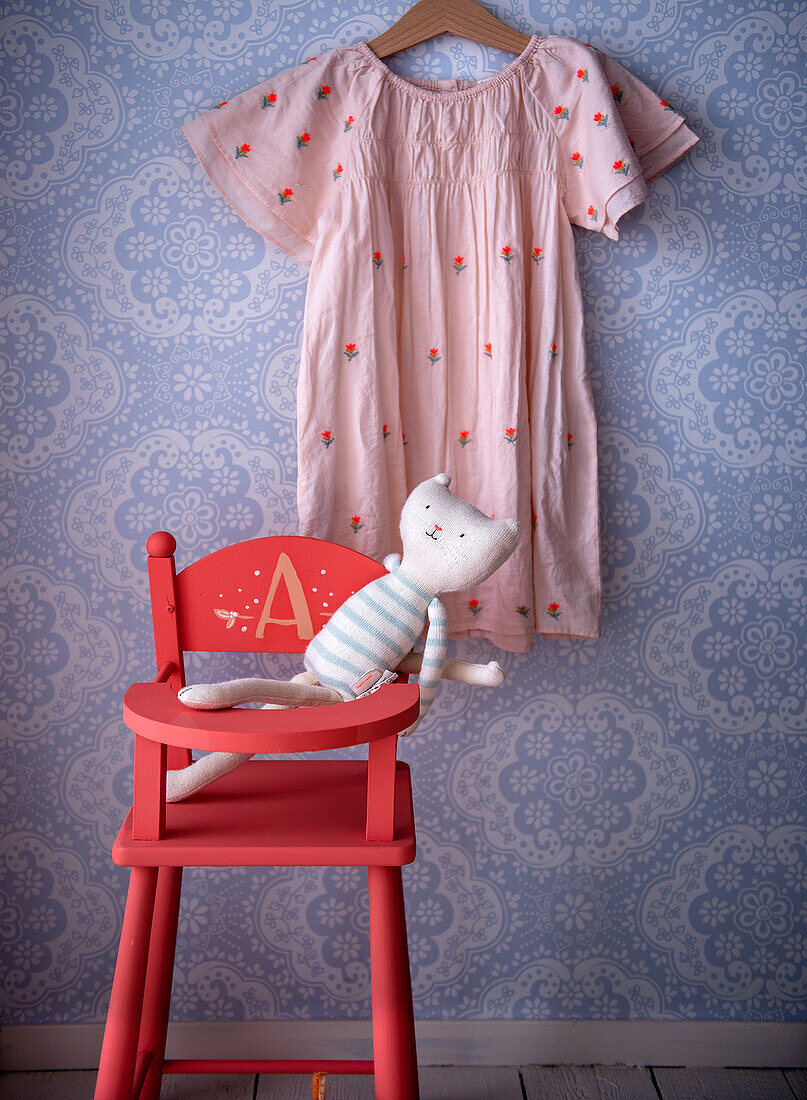 Hängendes rosa Kinderkleid mit Blumenmuster und Stoffkatze auf rotem Kinderstuhl