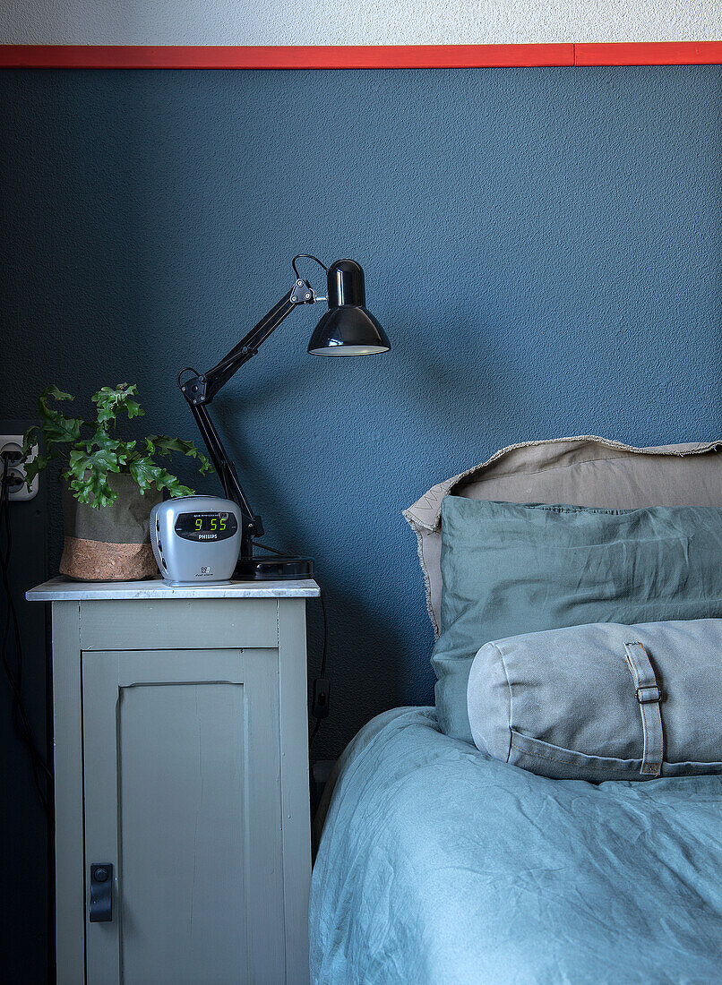 Bedside table with lamp and alarm clock next to bed with blue bed linen