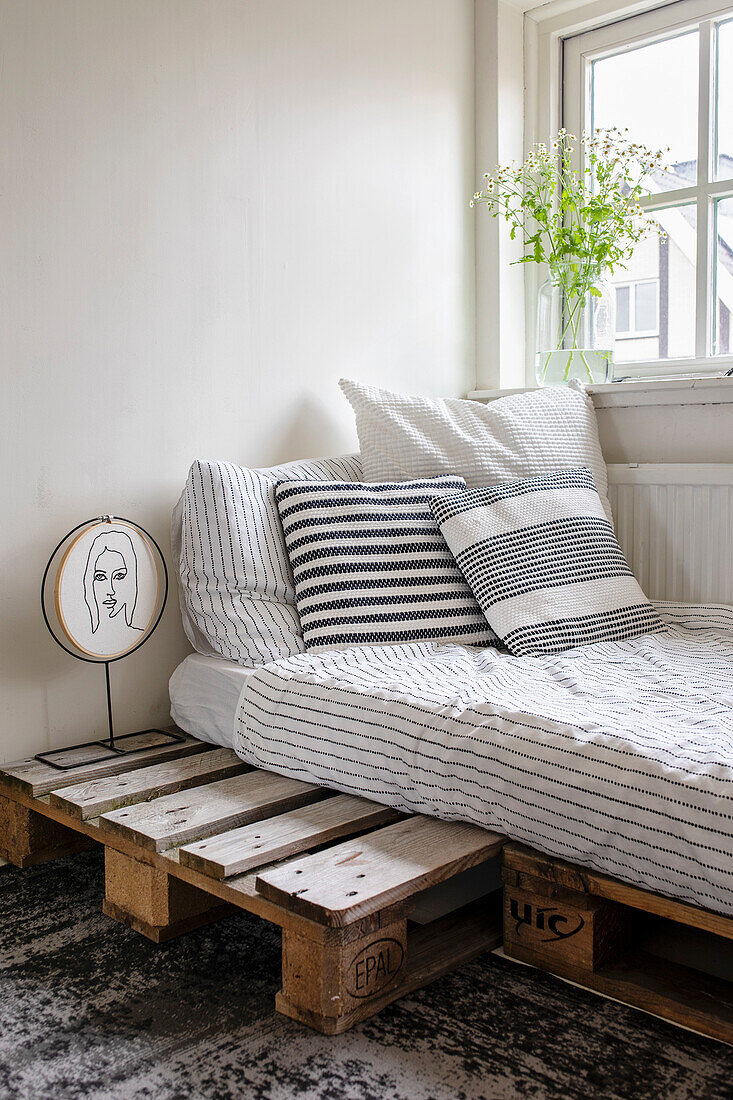 Bed on pallets with striped bed linen and decorative pillows next to window