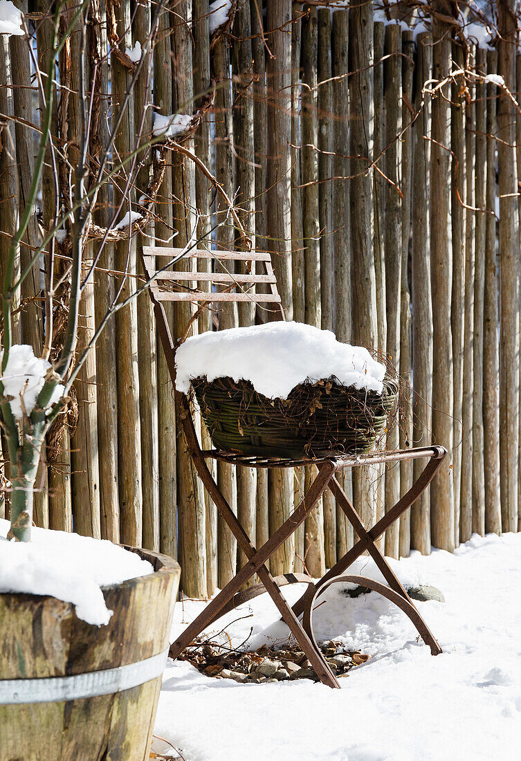 Schneebedeckter Weidenkorb auf Gartenstuhl vor Holzzaun im winterlichen Garten