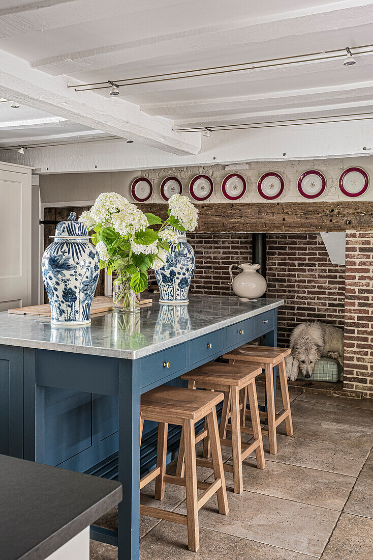 Country-style kitchen with blue cooking island and rustic fireplace, blue and white tea urns