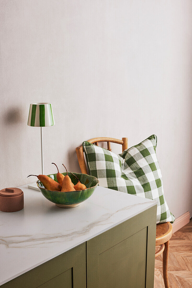 Green fruit bowl and lamp on sideboard next to green and white chequered cushion cover on wooden chair