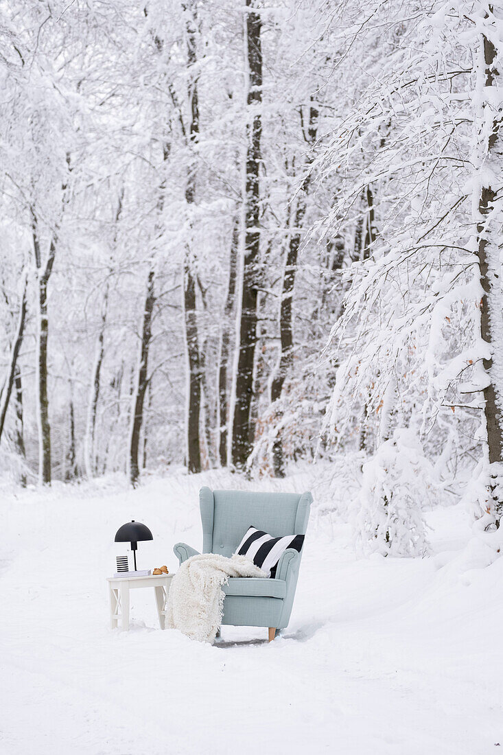 Hellblauer Ohrensessel mit Decke und Kissen in verschneitem Winterwald