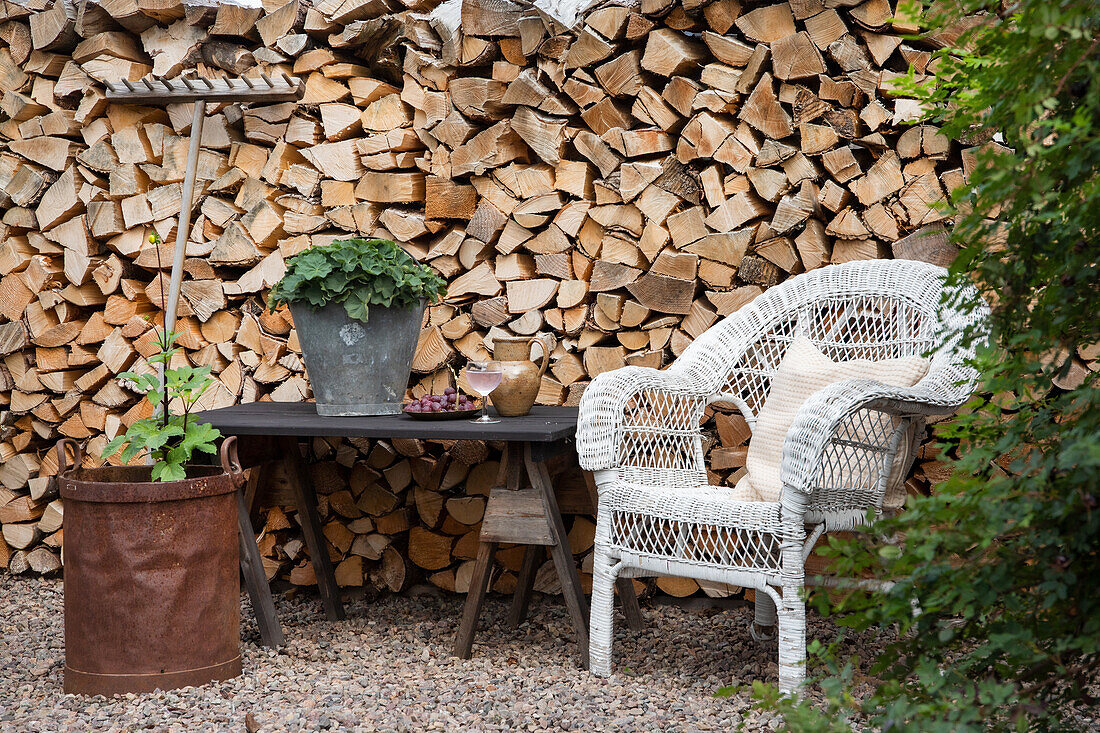 White rattan armchair and black table in front of pile of wood in the garden