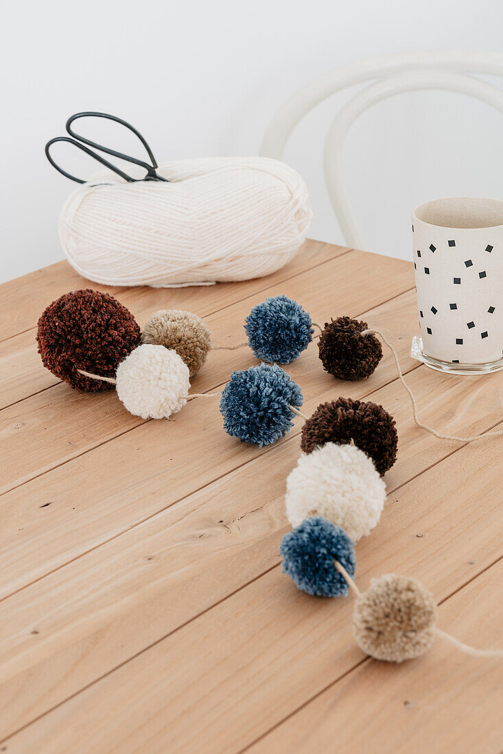 DIY garland made from wool pompoms, scissors and yarn on a wooden table