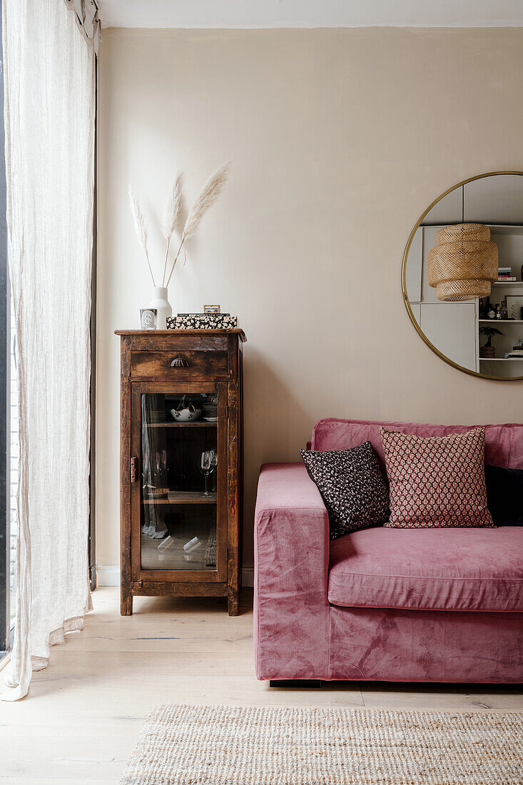 Pink sofa with patterned cushions next to rustic wooden cabinet and round wall mirror