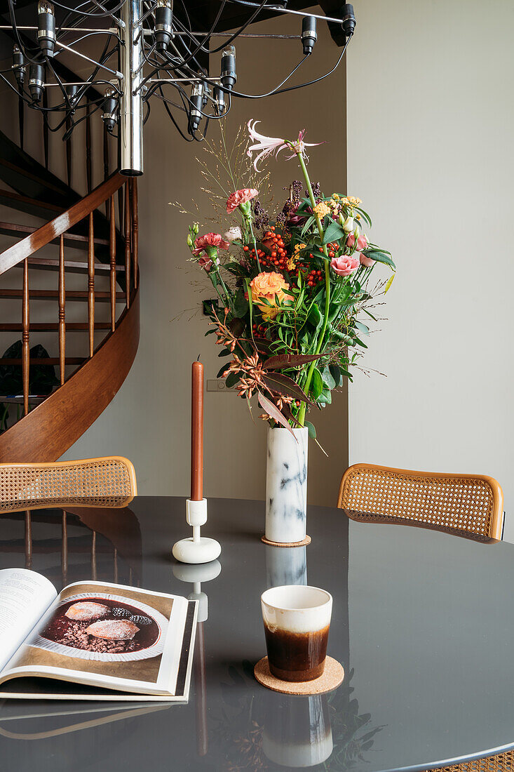 Round dining table with bouquet of flowers in marble vase, book and candle under modern hanging lamp