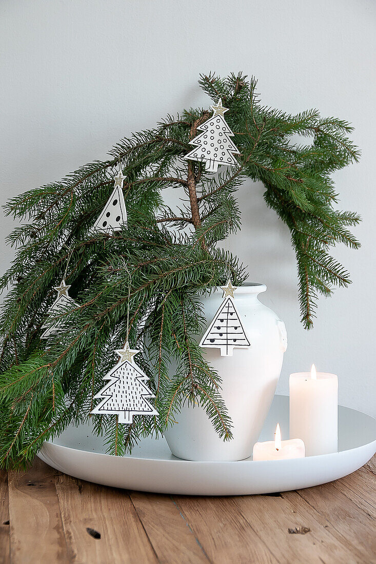 White vase with fir branches and Christmas tree pendants on a round tray with burning candles