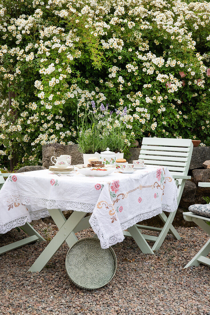 Gedeckter Gartentisch, Tischdecke mit Blumenstickerei und blühender Strauch