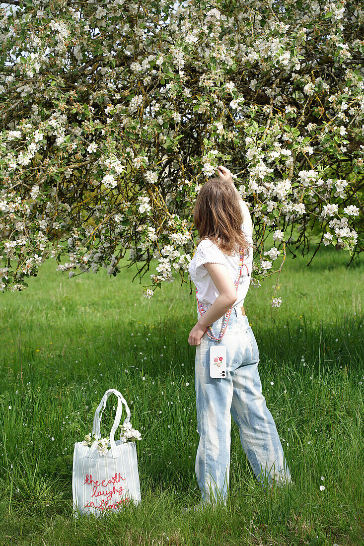 Blossoming fruit tree in the countryside, woman with smartphone and bag in the meadow