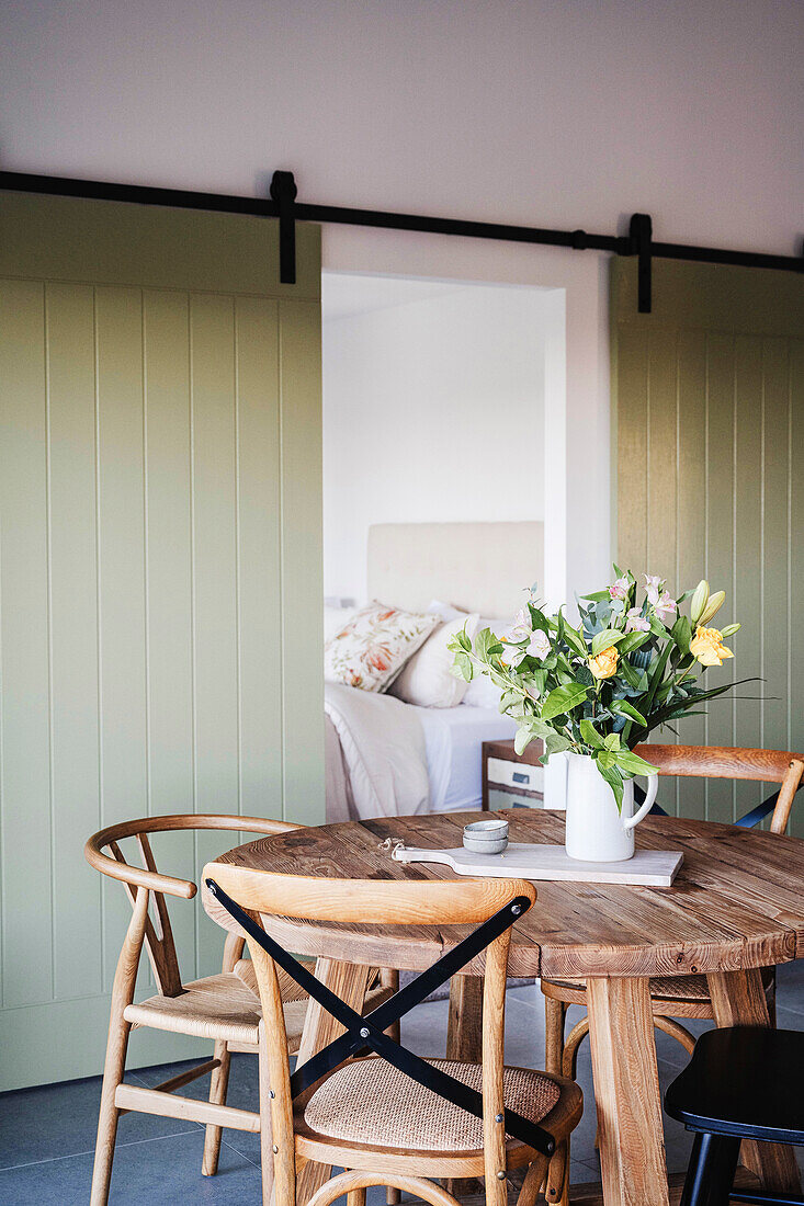 Round wooden table with bouquet of flowers in front of sliding door with view into bedroom