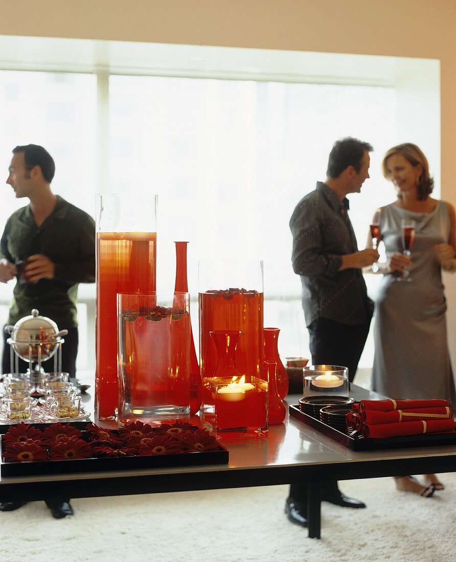 Tisch mit dekorativen roten Glasgefässen und Gerbera-Köpfen auf einem Tablett, im Hintergrund Partygäste