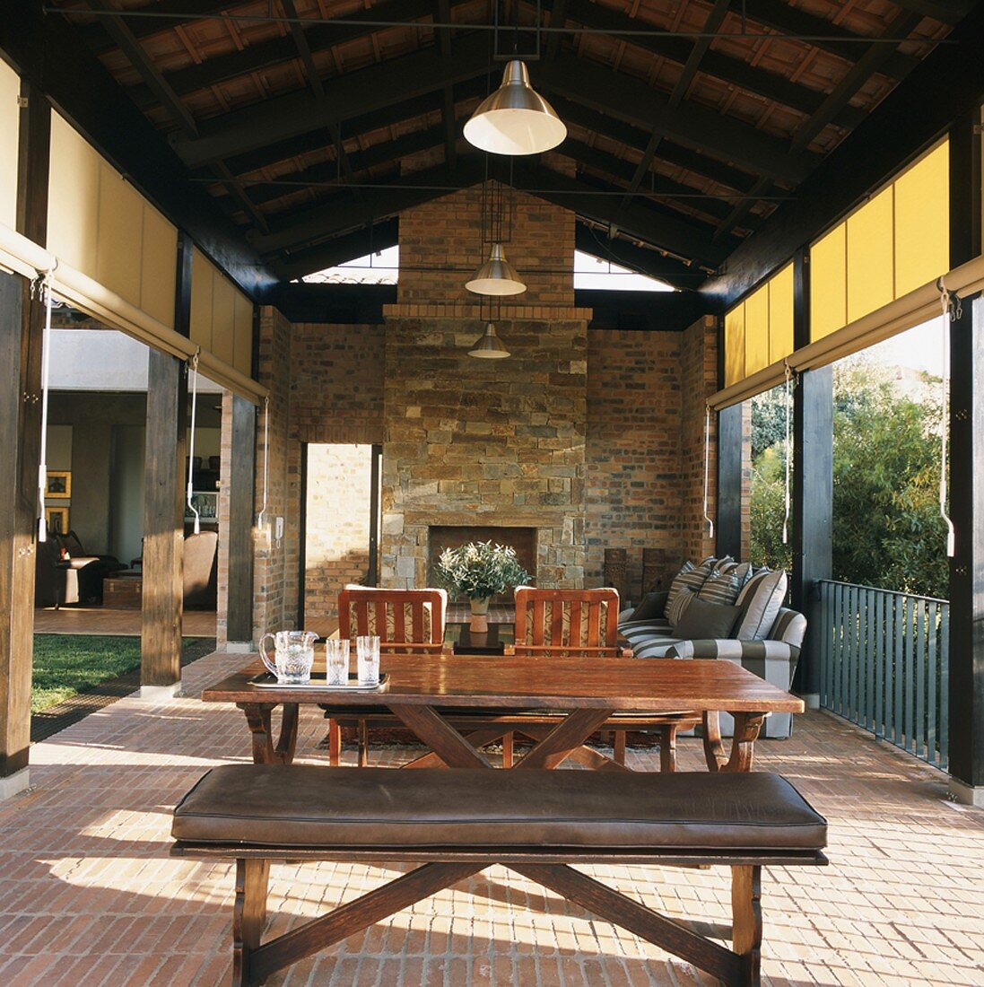 Wohn/Essraum im Freien - Terrasse mit rustikalem Kamin unter der offenen Dachkonstruktion und seitlichen Sonnenschutz-Rollos