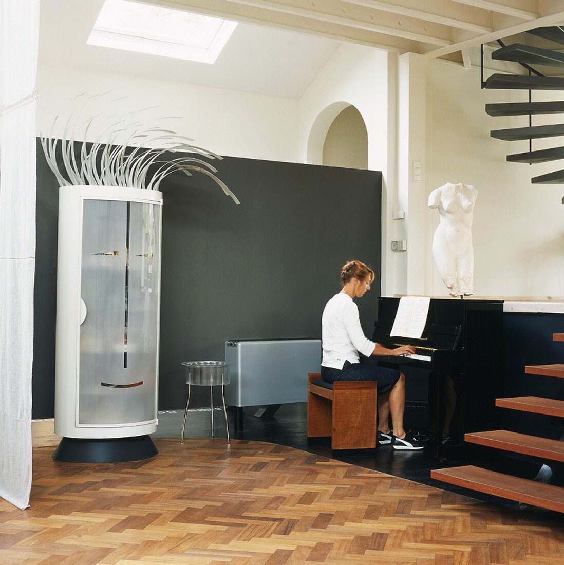 Whimsical cylindrical cupboard with stylised face and hair next to woman playing piano in loft apartment with open staircase