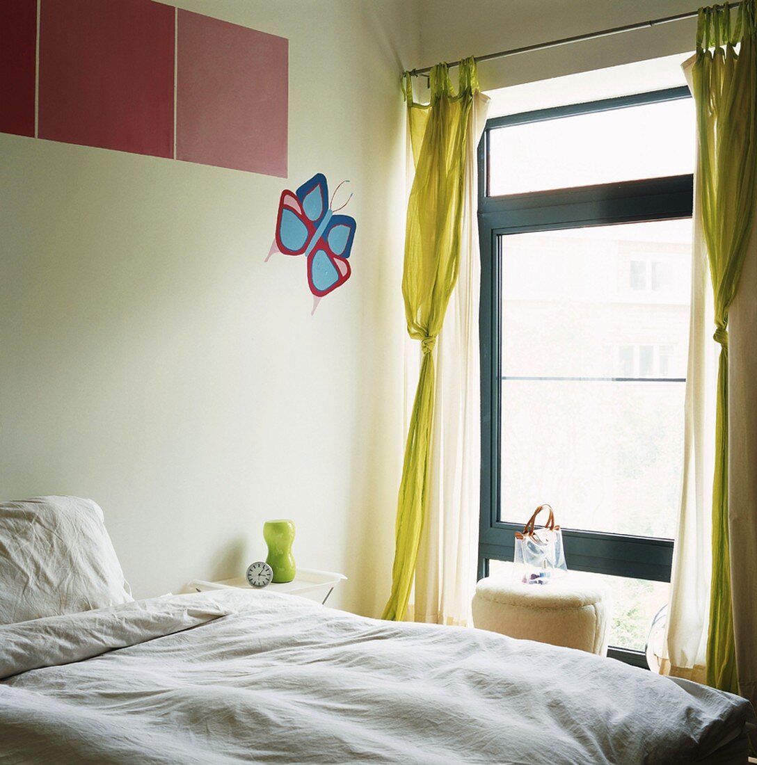 Bett vor einem Fenster mit gelben Vorhangschals und grafisch buntem Schmetterling an der Wand