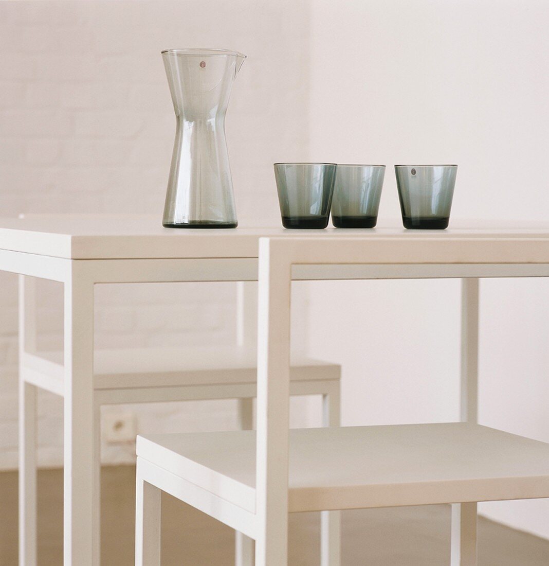 Karaffe und rauchgraue Gläser auf schlichtem, weißem Tisch mit zwei Stühlen