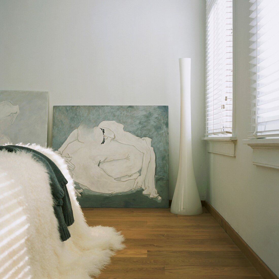 Weisser Schlafraum mit plüschiger Decke auf dem Bett, geschlossenen Jalousien und Akt-Malerei im Hintergrund