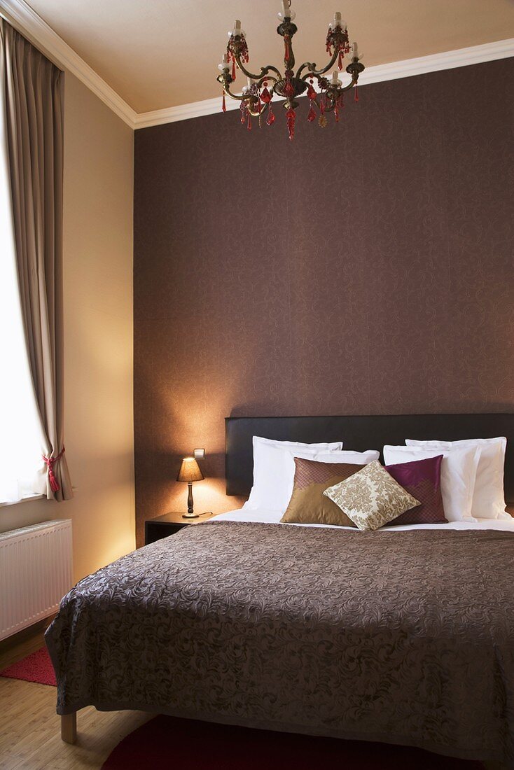 Einfacher Schlafraum mit brauner Wandtapete hinter dem Doppelbett und Kronleuchter mit roten Glaselementen
