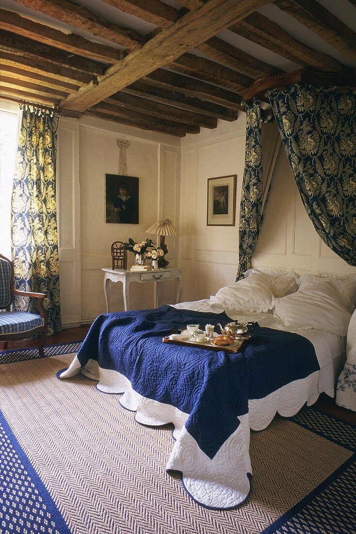 Bedroom with wood-beamed ceiling, canopy, rug and blue and white bedspread with breakfast tray on bed