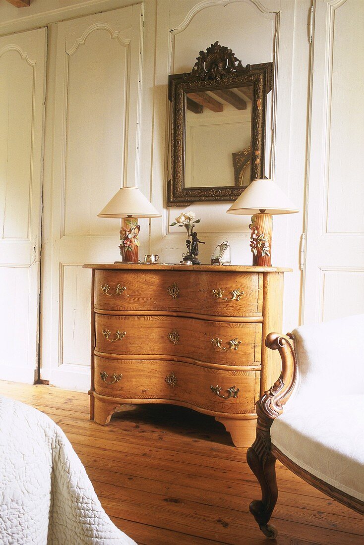 Table lamps on antique chest of drawers below antique mirror with carved wooden frame
