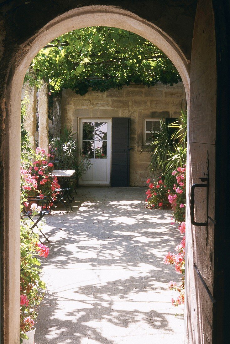 Blick durch einen Rundbogen auf sonnigen Innenhof mit schattenspendender Weinpergola und vielen Blumen