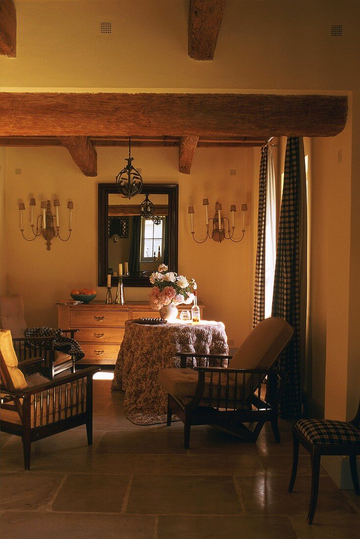 Cool, shaded interior with chest of drawers, framed mirror and wooden chairs with soft upholstery; round wooden table with floor-length cloth and elaborate bouquet