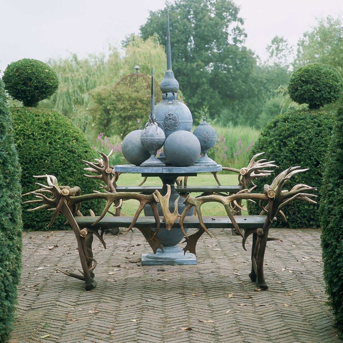 Zwei Sitzbänke mit ausgefallenen Tier-Geweih-Lehnen und einem runden Tisch mit dekorativen Metallkugeln; rundherum in Form geschnittene Hecken