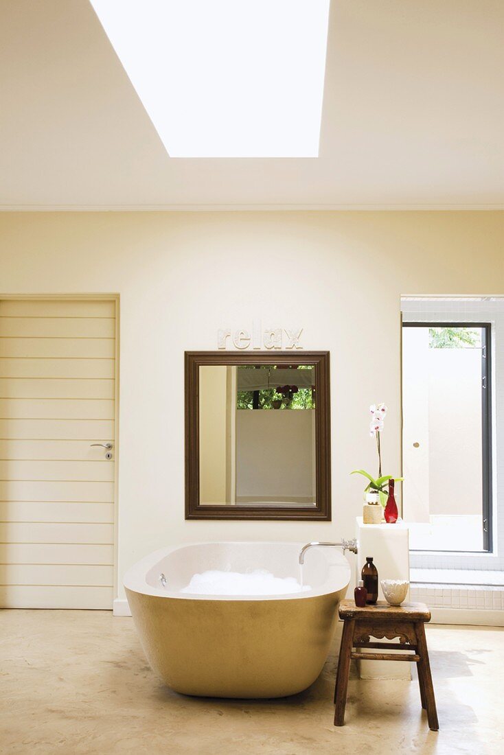 Freistehende Badewanne mit rechteckigem, holzgerahmtem Spiegel dahinter; daneben ein einfacher Holzhocker mit Badeutensilien