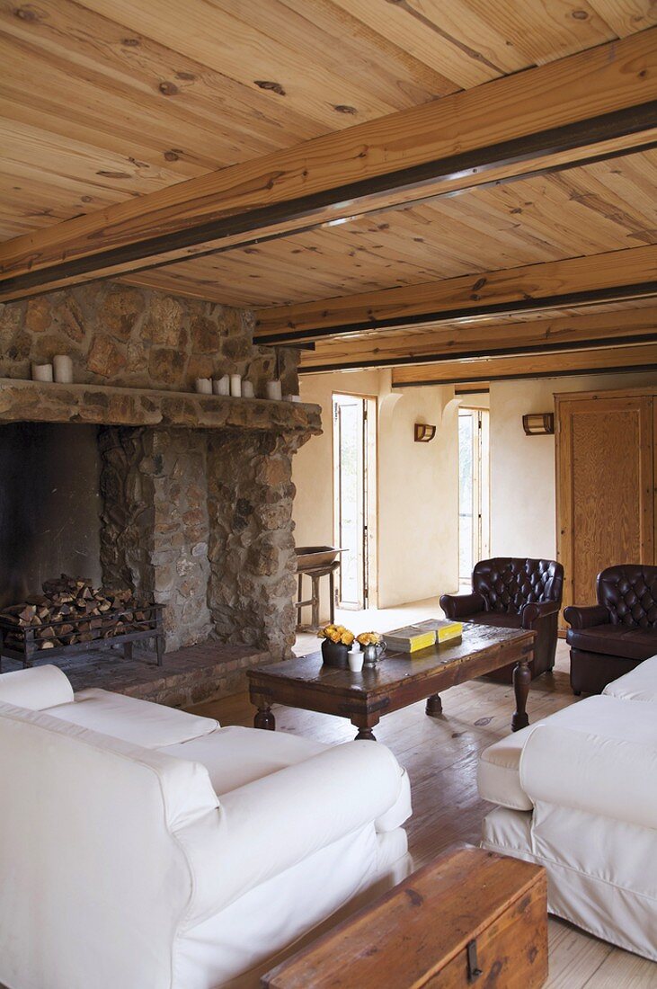 Herrschaftlicher Wohnraum mit großem, gemauertem Natursteinkamin, weissen Sofas und Ledersesseln unter einer niederen Holzdecke mit runden Balken