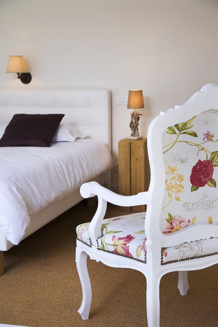 Bedroom with simple bed, rustic wooden bedside table and white chair with floral upholstery