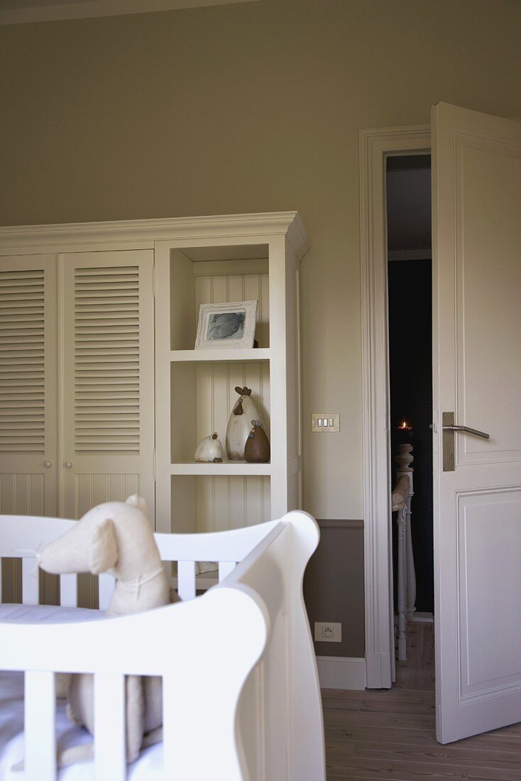 Weisser Kinderzimmerschrank mit Lamellentüren und Regalen; davor ein weisses Kinderbett mit Stoffhund