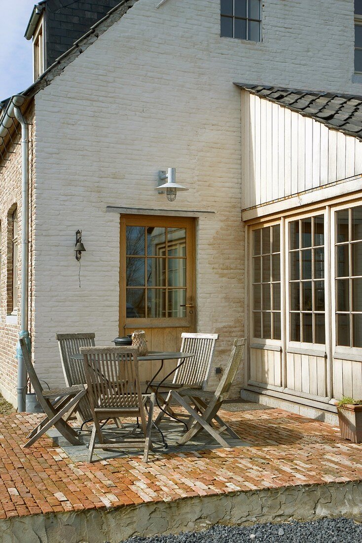 Backsteinhaus mit gepflasterter Sonnenterrasse; darauf einen runde Metalltisch und Klappstühle aus Holz