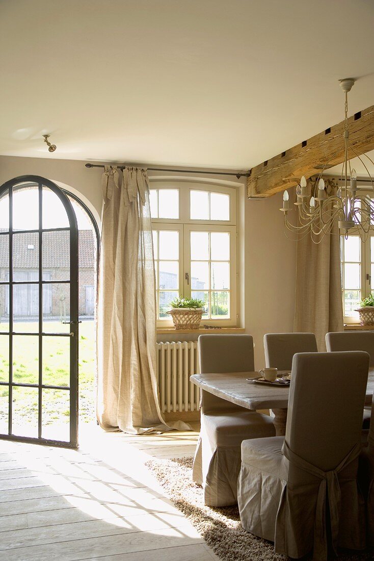 Ess- und Wohnraum mit großem Esstisch und Stühle mit eleganten Hussen; im Hintergrund die geöffnete Terrassentür aus Glas mit Metallrahmen
