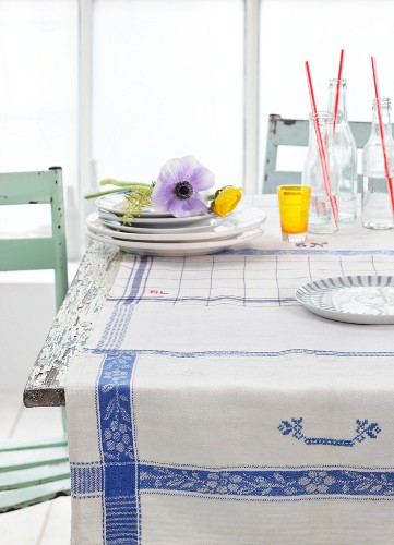 Blau-weiße Geschirrtücher als Tischdecke, Tellerstapel mit Anemonen und Flaschen auf rustikalem Küchentisch