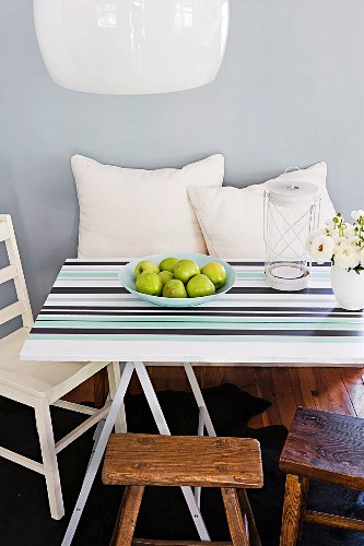 Mit unterschiedlichen Streifen gestaltete Tischplatte auf Holzböcken, rundum Sitzgelegenheiten und weiße Kissen vor pastellblauer Wand