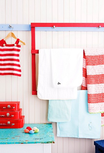 Rot gestrichener Holzrahmen mit Querleiste zum Aufhängen verschiedener Tücher im Kinderzimmer, daneben an der blauen Hakenleiste rotgestreiftes Kinderkleidchen