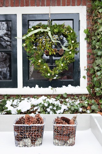 Fassadenausschnitt - Herzförmiger Kranz vor Fenster aufgehängt und Aufbewahrungskörbe mit Pinienzapfen im Schnee
